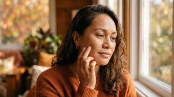 39-year-old Pacific Islander Australian woman with medium skin tone gently touching her cheek in warm autumn window light, examining her skin texture