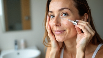 Woman's hands applying Medik8 Niacinamide Peptides serum to her cheek using pressing motion in natural bathroom lighting