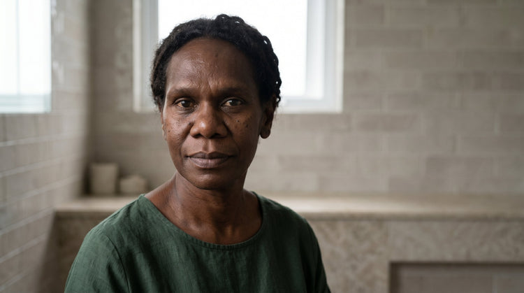 52-year-old Indigenous Australian non-binary person with dark brown skin holding a glass serum bottle near their face in soft natural light, calm expression in a minimal bathroom setting