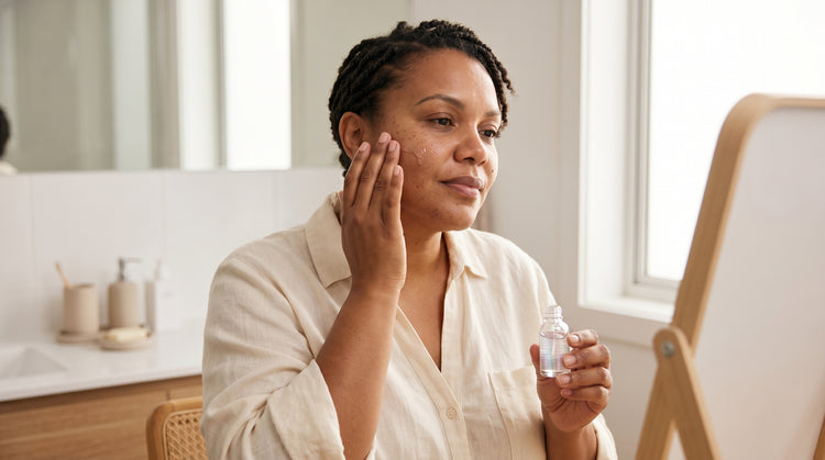 44-year-old African Australian woman with olive skin applying a serum to her cheek at a bathroom vanity, warm natural lighting highlighting her skin texture