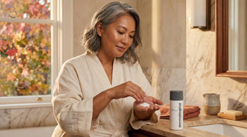 52-year-old East Asian Australian woman with medium skin tone applying serum to her cheek in a warmly lit bathroom, looking calm and confident