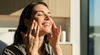 A woman gently massages moisturiser into her cheeks, eyes closed and smiling, bathed in warm sunlight — conveys a relaxed, post-care skincare moment.